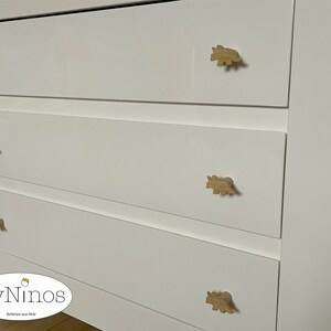 Op de afbeelding: Witte commode met vier lades en houten knoppen in de vorm van een dinosaurus. De commode heeft de tekst "ByNinos" en "Sch&ouml;nes aus Holz" op de bodem.