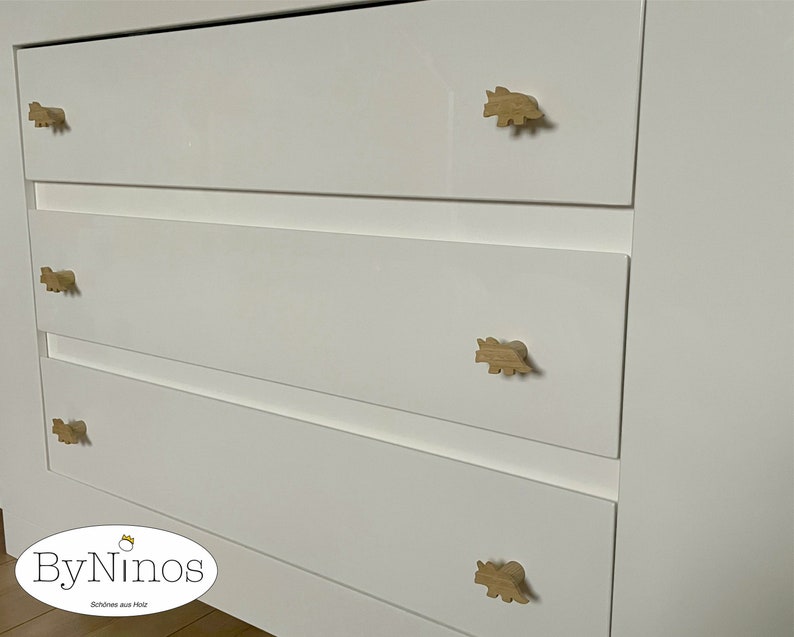 Op de afbeelding: Witte commode met vier lades en houten knoppen in de vorm van een dinosaurus. De commode heeft de tekst "ByNinos" en "Sch&ouml;nes aus Holz" op de bodem.