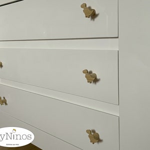 Op de afbeelding: Witte commode met vier lades en houten knoppen in de vorm van een dinosaurus. De commode heeft een logo met de tekst "ByNinos" en daaronder de tekst "Sch&ouml;nes aus Holz".