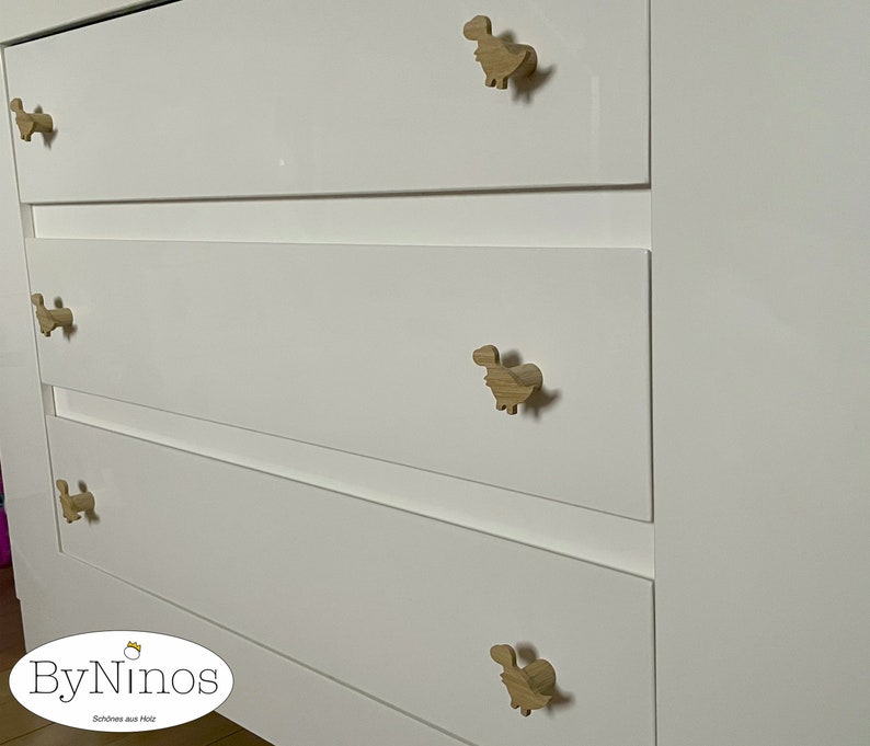 Op de afbeelding: Witte commode met vier lades en houten knoppen in de vorm van een dinosaurus. De commode heeft een logo met de tekst "ByNinos" en daaronder de tekst "Sch&ouml;nes aus Holz".