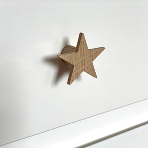 May include: A wooden star-shaped drawer pull with a light brown finish. The pull is mounted on a white surface.