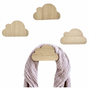 Puede incluir: Conjunto de cuatro ganchos de pared en forma de nube de madera. Los ganchos están hechos de madera natural y tienen un color marrón claro. Un paño rosa está colgado en uno de los ganchos.