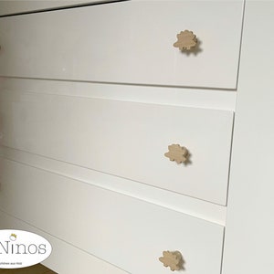 Op de afbeelding: Witte commode met vier lades en houten knoppen in de vorm van een wolk. De commode heeft een logo met de tekst "ByNinios" en "Sch&ouml;nes aus Holz".