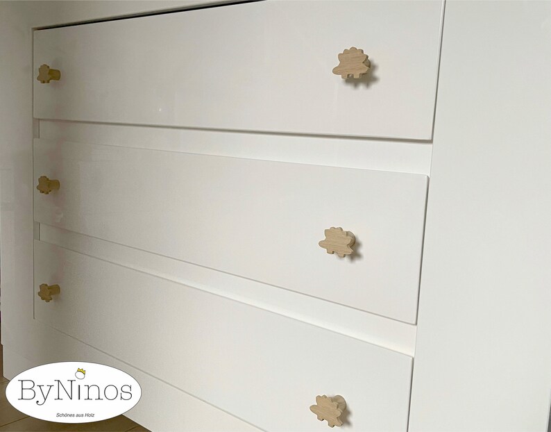 Op de afbeelding: Witte commode met vier lades en houten knoppen in de vorm van een wolk. De commode heeft een logo met de tekst "ByNinios" en "Sch&ouml;nes aus Holz".