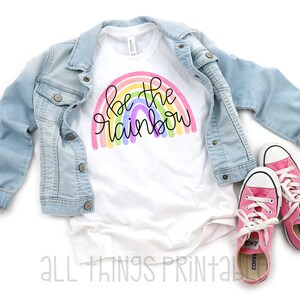 May include: White t-shirt with a rainbow design and the text "Be the rainbow" printed on it. The shirt is paired with a light blue denim jacket and pink sneakers.