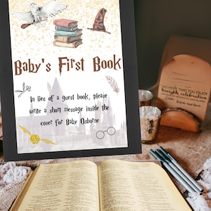 May include: A framed sign with the text "Baby's First Book" and illustrations of a snowy owl, books, and a sorting hat. The sign encourages guests to write a message for Baby Osborne. An open book and pens are in the foreground.