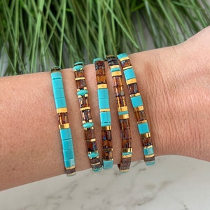 Peut inclure: Cinq bracelets empilables avec des carreaux turquoise et bruns et des accents dorés. Les bracelets sont empilés sur le poignet d'une personne.