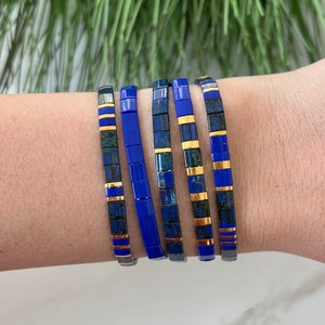 May include: Five stackable bracelets in a blue and gold color scheme. The bracelets are made of small square tiles with a blue background and gold accents. The bracelets are stacked on a person's wrist.