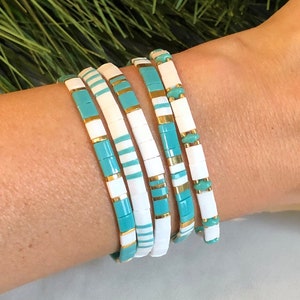Peut inclure: Ensemble de cinq bracelets en perles turquoise et blanches avec des accents dorés. Les bracelets sont superposés au poignet d'une personne.