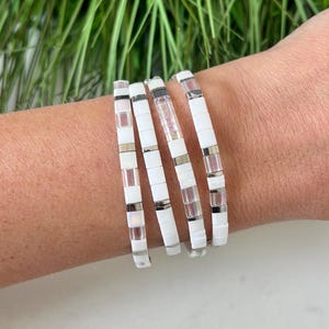 May include: Four white and silver beaded bracelets stacked on a wrist. The bracelets are made of small, square beads with a subtle iridescent sheen.