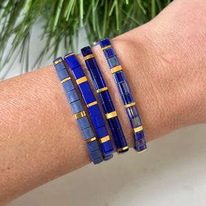 May include: Four blue and gold bracelets stacked on a wrist. The bracelets are made of small square beads with gold accents.