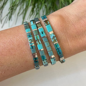 May include: Four turquoise and silver bracelets stacked on a wrist. The bracelets are made of small, square tiles with a turquoise and silver pattern.