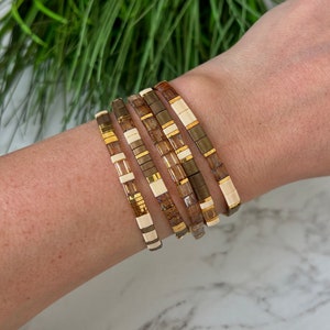 May include: A stack of five bracelets with brown, gold, and white beads. The bracelets are stacked on a person's wrist.