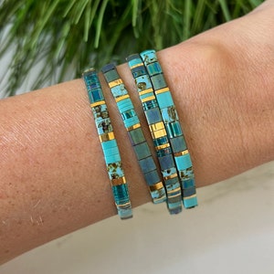 May include: Four turquoise and gold bracelets with iridescent accents. The bracelets are stacked on a person's wrist.