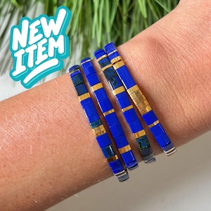 Puede incluir: Cuatro pulseras apilables de color azul oscuro con detalles dorados. Las pulseras están hechas de pequeñas baldosas cuadradas.