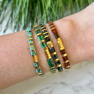 Könnte beinhalten: Vier goldene Armbänder mit unterschiedlichfarbigen Fliesen. Die Armbänder sind auf dem Handgelenk einer Person gestapelt. Die Fliesen sind türkis, grün, schwarz und orange.