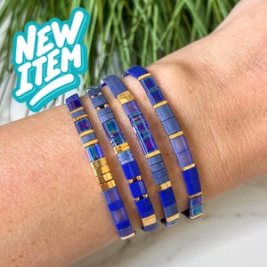 Puede incluir: Se muestran cinco pulseras azules y doradas en una muñeca. Las pulseras están hechas de pequeñas cuentas rectangulares en tonos azules y dorados. Las pulseras están etiquetadas como "NEW ITEM" en una burbuja turquesa.