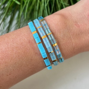 Puede incluir: Tres pulseras apiladas con detalles en azul claro y dorado. Las pulseras están hechas de pequeñas cuentas cuadradas.