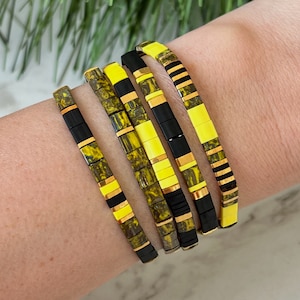 May include: A set of five bracelets with yellow and green patterned beads. The bracelets are stacked on a person's wrist.