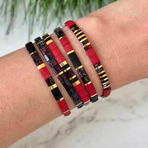 May include: Five stacked bracelets with red, black, and gold beads. The bracelets are made of small, square beads.