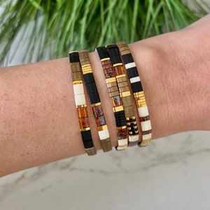 Puede incluir: Un conjunto de seis pulseras coloridas con cuentas negras, blancas, doradas y marrones. Las pulseras están hechas de diferentes materiales, incluyendo cerámica, metal y vidrio.