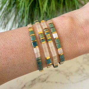May include: Stack of four bracelets in a teal and gold colour scheme. The bracelets are made of small square tiles with a glossy finish.