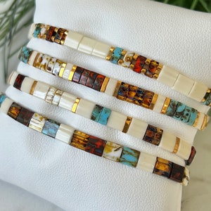 May include: Stack of five bracelets with white, brown, orange, and turquoise beads. The beads are arranged in a geometric pattern and have a gold accent.