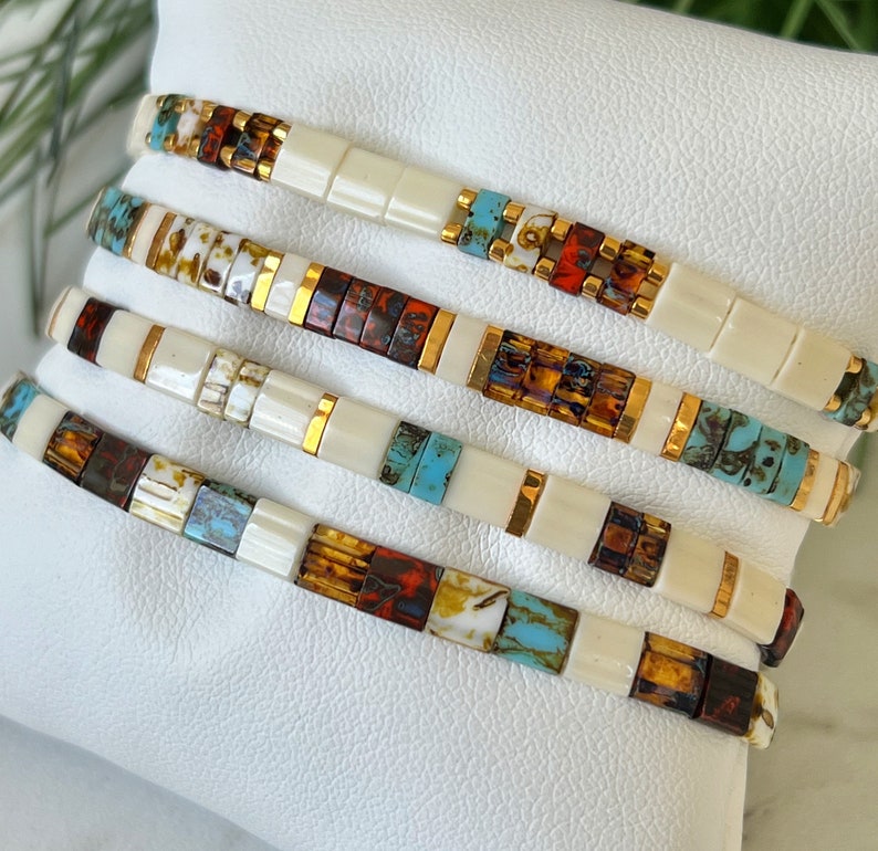 May include: Stack of five bracelets with white, brown, orange, and turquoise beads. The beads are arranged in a geometric pattern and have a gold accent.