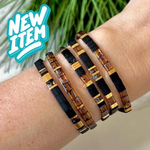 May include: Six bracelets with a mix of black, gold, and iridescent beads are displayed. The bracelets have a flat, rectangular design. The text "NEW ITEM" is in a teal bubble in the upper left corner. The bracelets are on a person's arm.
