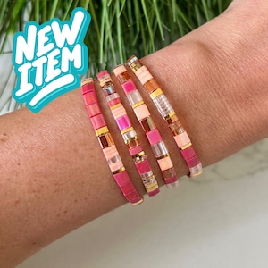 Può includere: Un set di cinque braccialetti colorati con perline in tonalità rosa, arancio e oro. I braccialetti sono impilati sul polso di una persona.