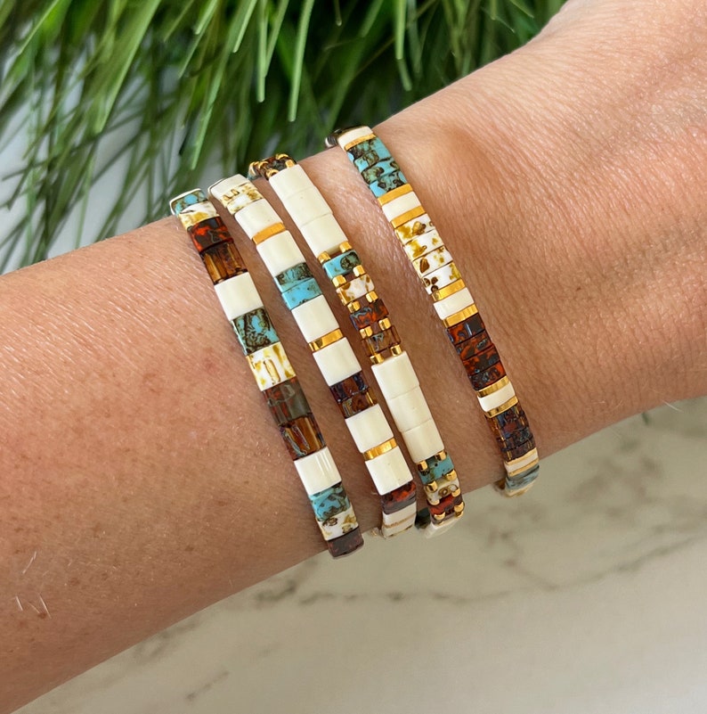 May include: Four stacked bracelets with a white base and colorful patterns. The bracelets feature turquoise, brown, and gold accents.