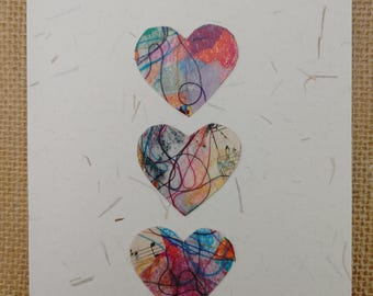 May include: A handmade greeting card with three heart-shaped cutouts. Each heart features a colorful abstract design with blue, pink, and orange hues. The card is made of textured white paper.