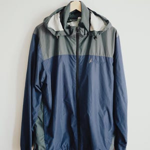 K&ouml;nnte beinhalten: Eine blau-graue Kapuzenjacke mit durchgehendem Rei&szlig;verschluss, die an einem Holzb&uuml;gel h&auml;ngt. Die Jacke hat einen Kordelzug am Saum und ein kleines Logo auf der Brust. Die Jacke ist aus leichtem Material.