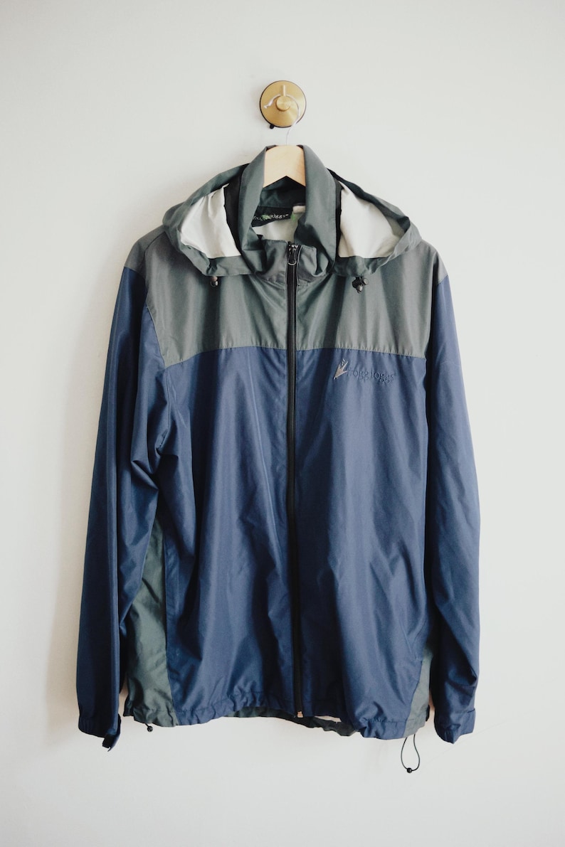 K&ouml;nnte beinhalten: Eine blau-graue Kapuzenjacke mit durchgehendem Rei&szlig;verschluss, die an einem Holzb&uuml;gel h&auml;ngt. Die Jacke hat einen Kordelzug am Saum und ein kleines Logo auf der Brust. Die Jacke ist aus leichtem Material.