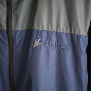 K&ouml;nnte beinhalten: Zweifarbige Jacke in Blau und Grau mit durchgehendem Rei&szlig;verschluss und Kapuze. Auf der Brust ist das gestickte Logo "frogg toggs" zu sehen. Das Material scheint ein leichter, wasserabweisender Stoff zu sein.