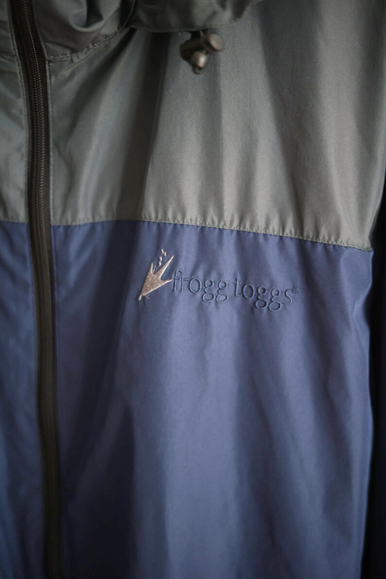 K&ouml;nnte beinhalten: Zweifarbige Jacke in Blau und Grau mit durchgehendem Rei&szlig;verschluss und Kapuze. Auf der Brust ist das gestickte Logo "frogg toggs" zu sehen. Das Material scheint ein leichter, wasserabweisender Stoff zu sein.
