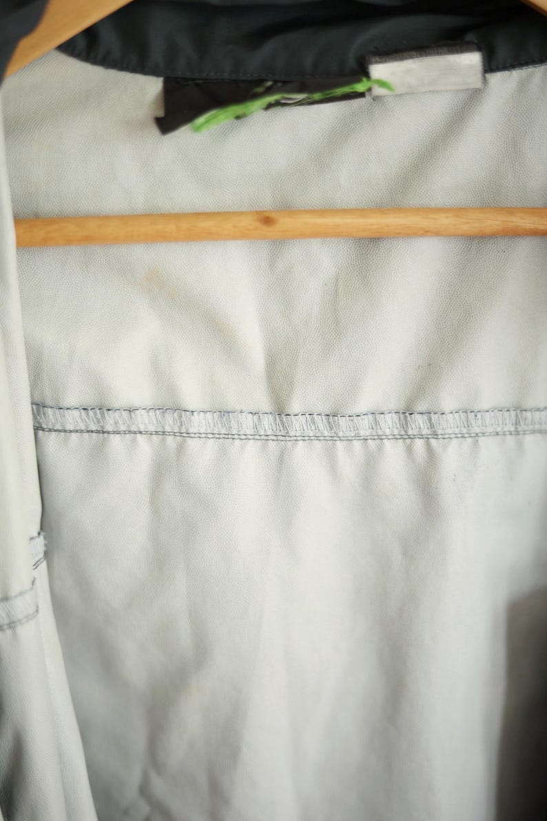 K&ouml;nnte beinhalten: Nahaufnahme des Innenraums einer hellfarbigen Jacke. Die Jacke h&auml;ngt an einem Holzb&uuml;gel. Der Innenstoff ist hellgrau mit einer horizontalen Naht aus grauen N&auml;hten. Die Oberseite der Jacke ist schwarz.