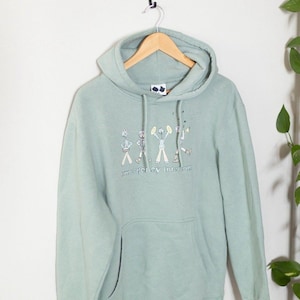 May include: A light green hoodie with a drawstring hood and a front pocket. The hoodie features a graphic of stick figures playing instruments, with the text "emergency interior" below the figures. The hoodie is hanging on a wooden hanger.