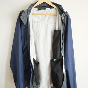 K&ouml;nnte beinhalten: Eine marineblaue und graue Kapuzenjacke mit hellgrauem Innenfutter, die an einem Holzb&uuml;gel h&auml;ngt. Die Jacke hat mehrere schwarze Taschen und einen Kordelzug am unteren Saum. Die Jacke wird vor wei&szlig;em Hintergrund pr&auml;sentiert.