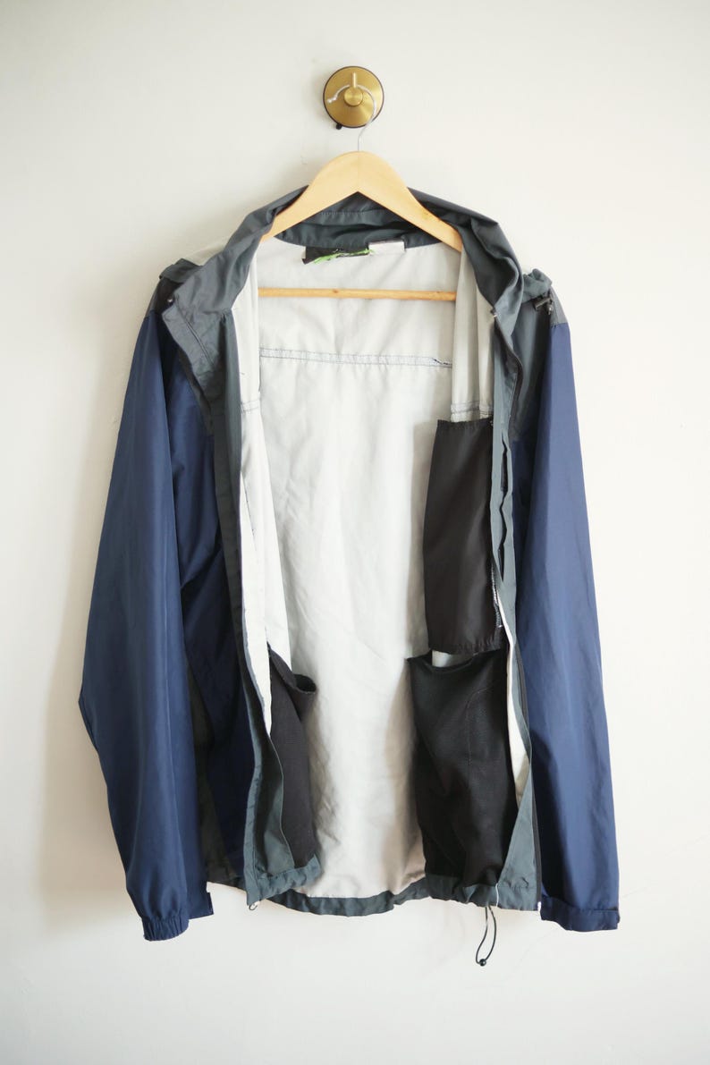 K&ouml;nnte beinhalten: Eine marineblaue und graue Kapuzenjacke mit hellgrauem Innenfutter, die an einem Holzb&uuml;gel h&auml;ngt. Die Jacke hat mehrere schwarze Taschen und einen Kordelzug am unteren Saum. Die Jacke wird vor wei&szlig;em Hintergrund pr&auml;sentiert.