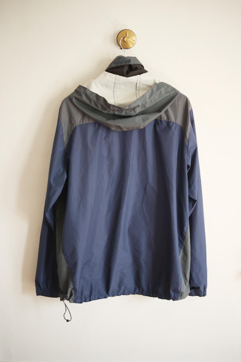 K&ouml;nnte beinhalten: Eine marineblaue und graue Kapuzenjacke h&auml;ngt an einer Wand. Die Jacke hat einen Kordelzug am unteren Saum und eine graue Kapuze. Die Oberseite der Jacke ist wei&szlig; und schwarz.