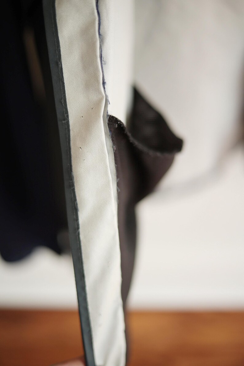 K&ouml;nnte beinhalten: Nahaufnahme eines Stoffabschnitts mit einem wei&szlig;en, strukturierten Panel, das von einer dunkelgrauen Umrandung begrenzt wird. Der Stoff scheint geschichtet zu sein, mit einem dunkelblauen Material auf der linken Seite.