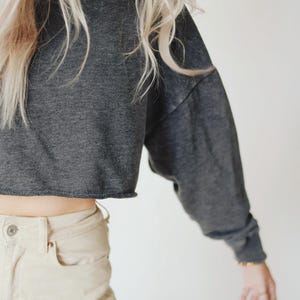 May include: A dark gray cropped sweatshirt with long sleeves and a raw hem. The sweatshirt is paired with light beige pants. The model has long blonde hair.