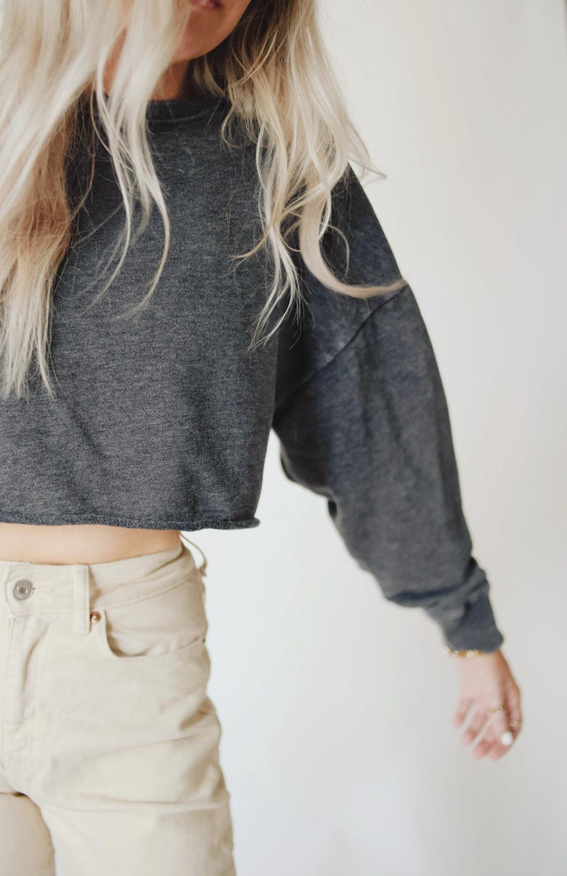 May include: A dark gray cropped sweatshirt with long sleeves and a raw hem. The sweatshirt is paired with light beige pants. The model has long blonde hair.