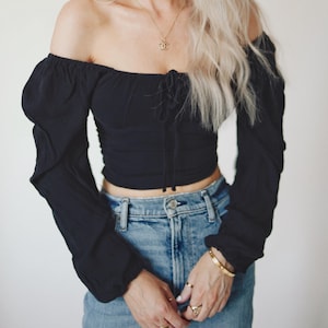 May include: Black off-the-shoulder top with long sleeves and a tie-up front. The top is paired with light-wash blue jeans. A gold necklace and bracelets are visible.