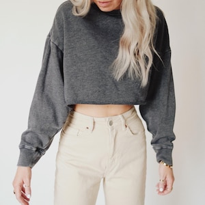May include: A charcoal gray cropped sweatshirt with long sleeves and a raw hem. Paired with light beige high-waisted jeans. The outfit is casual and comfortable, suitable for everyday wear.
