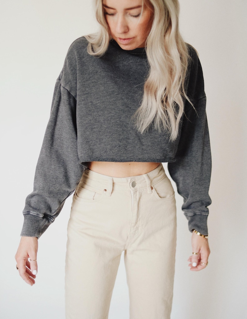 May include: A charcoal gray cropped sweatshirt with long sleeves and a raw hem. Paired with light beige high-waisted jeans. The outfit is casual and comfortable, suitable for everyday wear.