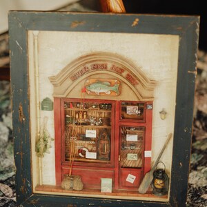 May include: A framed, three-dimensional diorama of a fishing shop, with a weathered blue frame. The shop has a red facade with a sign that reads "River's Edge Bait Shop" and a green fish emblem. Fishing rods, lures, and other items are visible inside.