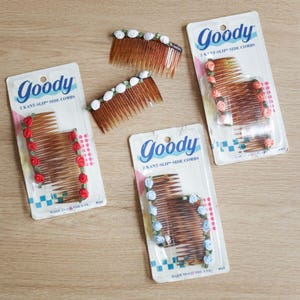 May include: Three packages of Goody 2 Kant-Slip side combs, each containing two amber-colored plastic combs. The combs are decorated with small fabric roses in red, light blue, and peach. Two loose combs with white rose accents are also included.