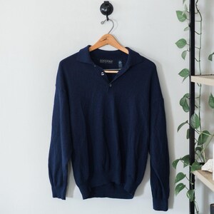 May include: A navy blue sweater with a collared neckline and three buttons. The sweater is hanging on a wooden hanger against a white wall. The brand name "Giasone" is visible on the label.
